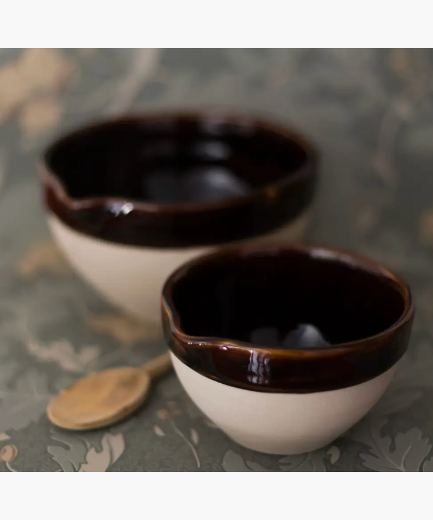 Two ceramic bowls with dark brown interiors and creamy exteriors sit on a floral patterned surface. A wooden spoon lies beside them, creating a rustic vibe. | Wildwood Home Co.